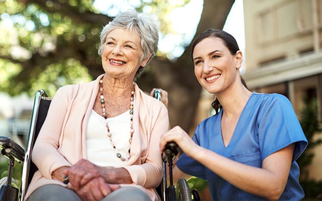 An elderly woman in a wheelchair smiles alongside a younger woman in blue scrubs outdoors.