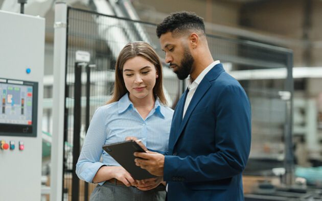 Two people in business attire discuss something on a tablet in a factory setting.