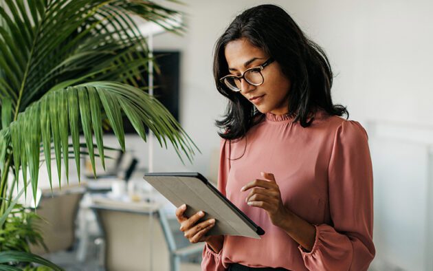 A woman in a pink blouse stands near a plant, looking at a tablet in her hand.