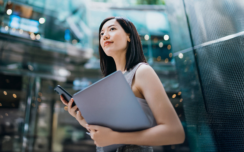 A woman holding a laptop and smartphone stands in a modern glass building, looking upwards with a neutral expression.