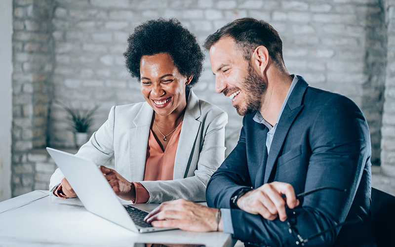 Two business professionals, a woman and a man, sitting at a desk, smiling, and looking at a laptop screen in a modern office setting.