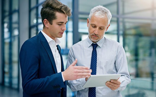 Two men in business attire stand indoors, discussing something on a digital tablet. One gestures while the other holds and looks at the tablet.