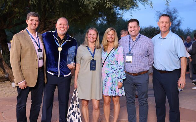 Six people stand outdoors posing for a group photo, all wearing name badges, with one person holding a glass award. Trees and other people are visible in the background.
