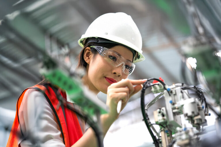Woman wearing a white hard hat, safety glasses, and orange vest inspects or works on machinery components in an industrial or manufacturing setting.
