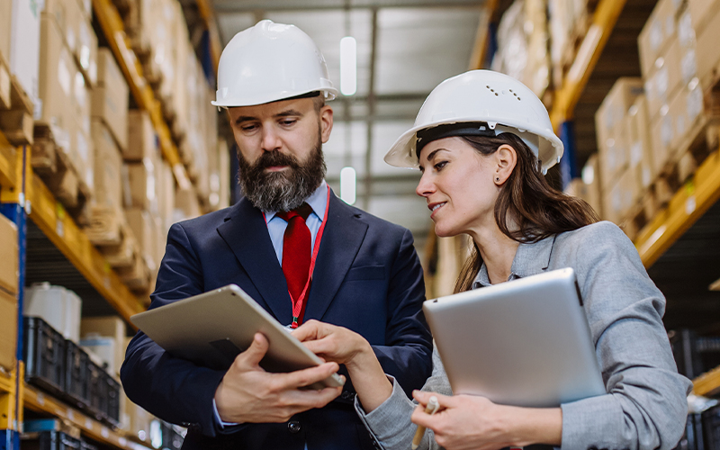 Two people wearing hard hats and business attire review information on a tablet in a warehouse with shelves stocked in the background.