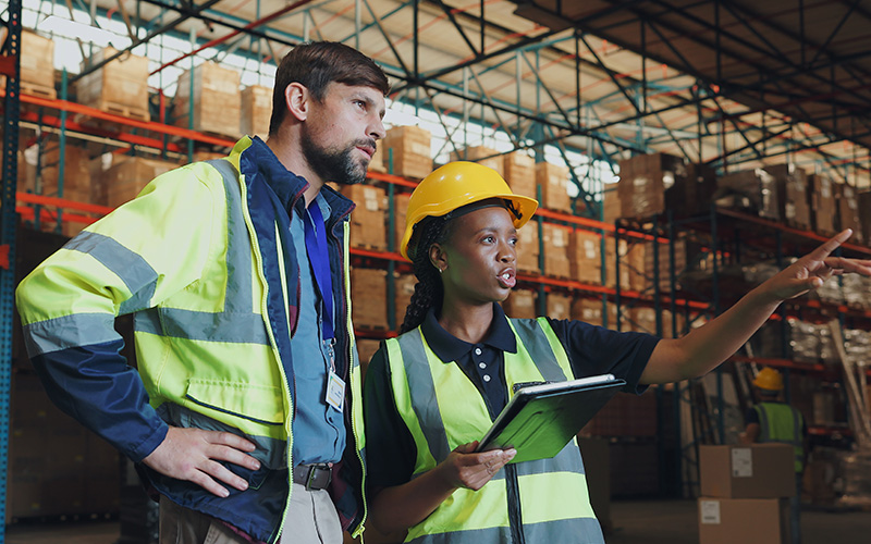 Two warehouse workers wearing safety vests and helmets stand in a storage facility; one holds a clipboard and points while the other looks in the indicated direction.