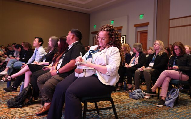 People sitting in rows at a conference or seminar, facing forward, with one woman in the foreground taking notes on a notepad.