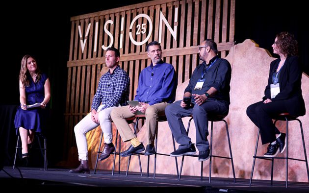 Five people sit on stools on a stage, engaged in a panel discussion with a wooden "VISION 25" sign in the background.