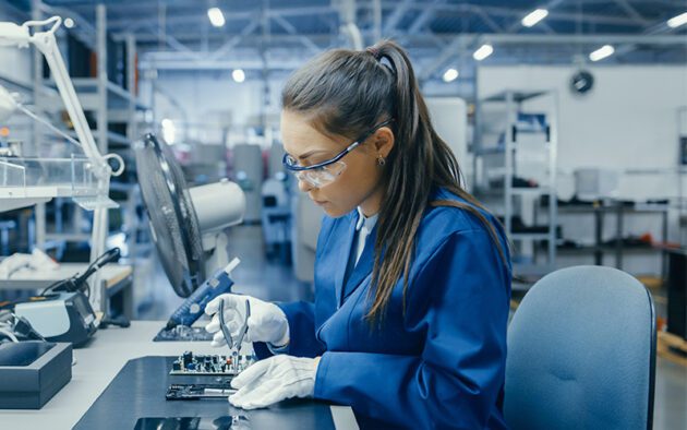 A woman wearing safety glasses and gloves works on electronic components with tools at a workstation in a laboratory or manufacturing facility.