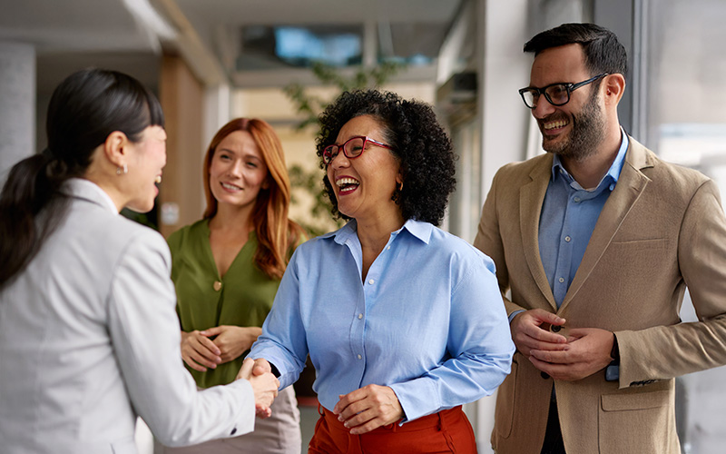 Four professionals stand indoors; two women are shaking hands while the other two colleagues smile and observe the interaction.