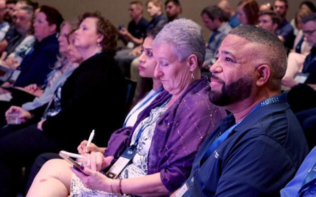 A group of people sit closely together in an audience at a conference, some taking notes and listening attentively to a speaker off camera.