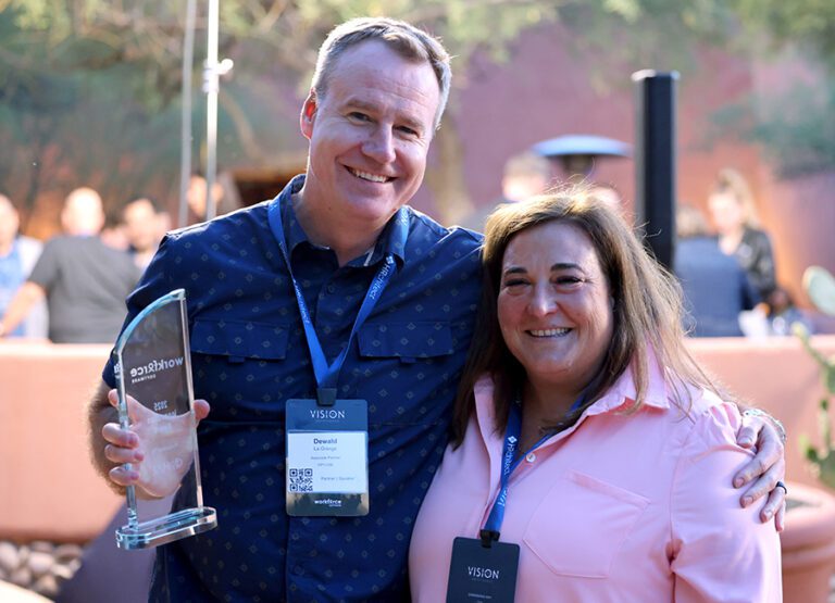 A man holding a glass award and a woman stand together outdoors, both wearing conference lanyards and smiling at the camera.