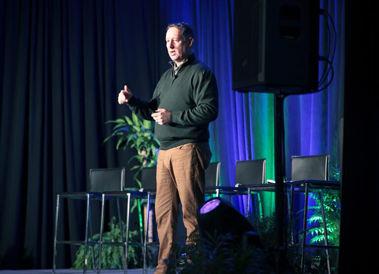 A man stands on stage giving a presentation, with empty chairs, plants, and stage lighting in the background.