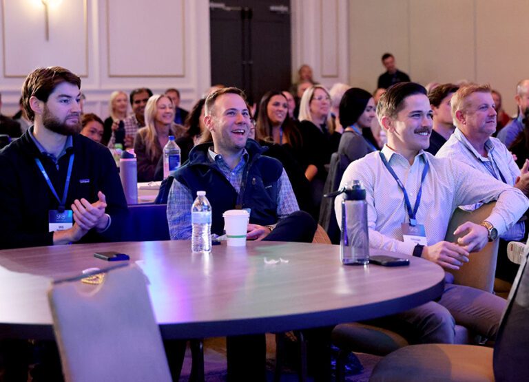 A group of people, mostly men, sit at round tables and watch a presentation in a conference room, some smiling and clapping.