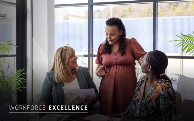 Three women are having a discussion at a conference table in a modern office, with documents in hand and sunlight coming through large windows. The text "Workforce Excellence" is displayed.