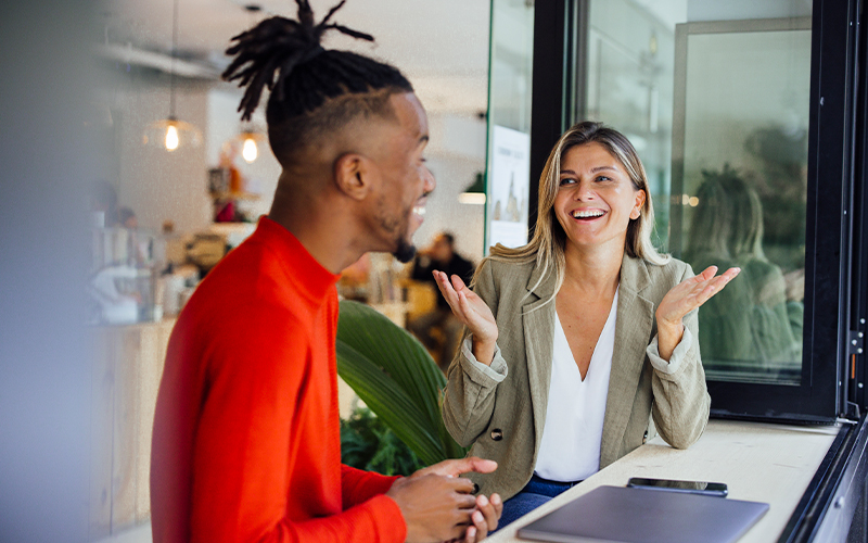 Two people sit by a window in a café, having a lively conversation. One person gestures with hands raised while the other listens and smiles. A laptop is on the counter beside them.