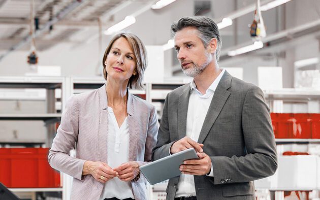 Two professionally dressed adults stand indoors; the man holds a tablet and gestures while the woman listens. Industrial shelving and bright lighting are visible in the background.
