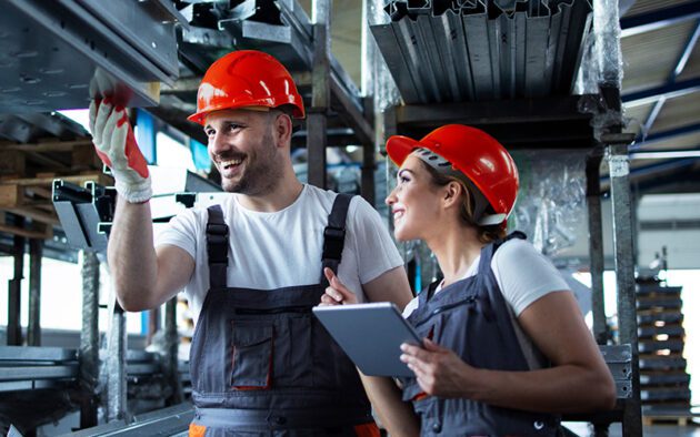 Two workers in overalls and orange hard hats stand in an industrial warehouse; one gestures toward metal shelves while the other holds a tablet and observes.
