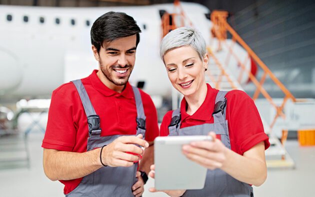Two aviation technicians in red shirts and gray overalls look at a tablet together in an aircraft hangar with a plane in the background.
