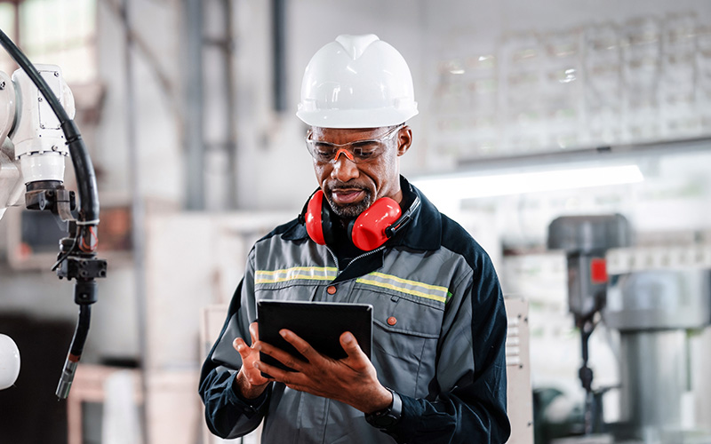 A man wearing safety gear, including a hard hat, safety glasses, and ear protection, uses a tablet in an industrial setting.