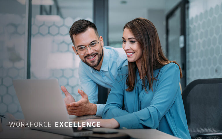 Two colleagues in blue shirts are working together at a desk with a laptop, smiling and discussing something on the screen. The text "WORKFORCE EXCELLENCE" appears in the corner.