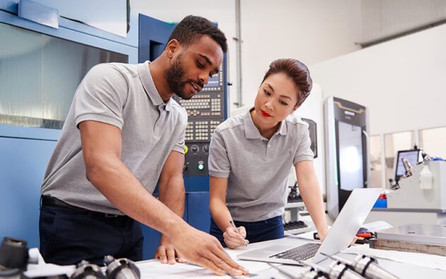 Two engineers in gray polo shirts review documents and work on a laptop beside industrial machinery in a modern manufacturing facility.