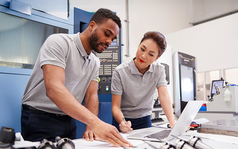 Two engineers in gray polo shirts review documents and work on a laptop beside industrial machinery in a modern manufacturing facility.