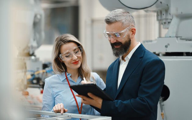 Two people in safety glasses and business attire review notes on a clipboard in an industrial or manufacturing setting with machinery in the background.