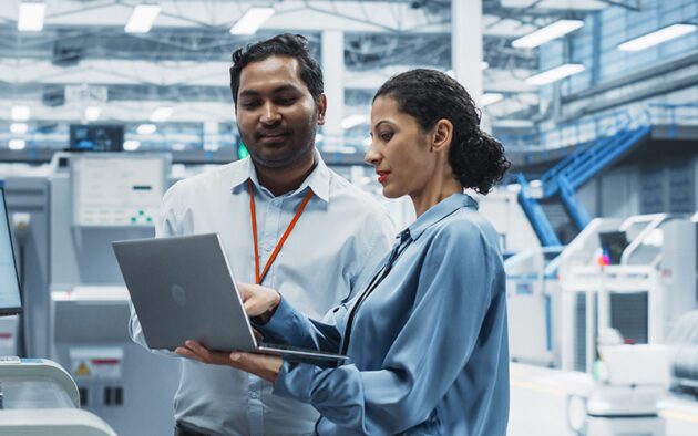Two people in business attire discuss information on a laptop in a modern industrial facility, with machinery and equipment visible in the background.