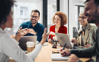 Five people sit around a table in an office, engaged in a lively discussion with laptops, notebooks, and coffee cups in front of them.