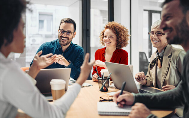 Five people sit around a table in an office, engaged in a lively discussion with laptops, notebooks, and coffee cups in front of them.