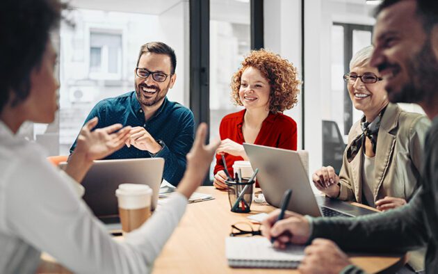 Five people sit around a table in an office, engaged in a discussion with laptops, notebooks, and coffee cups in front of them.