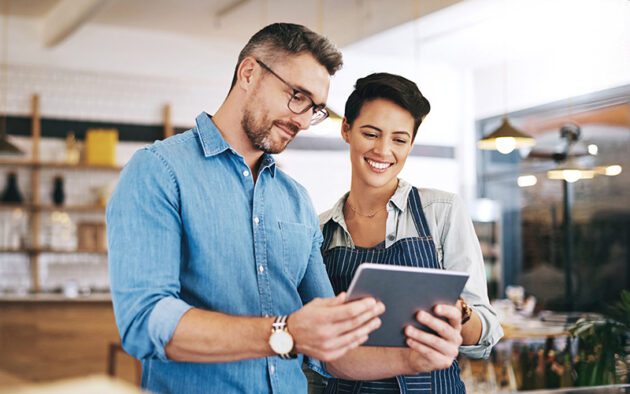 Two people standing together indoors, looking at a digital tablet and smiling; one wears glasses and a denim shirt, the other wears an apron over a shirt.