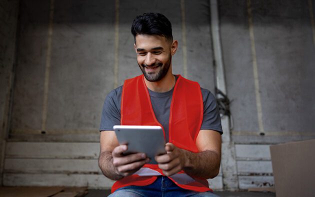 A man wearing a red safety vest sits indoors, smiling while using a tablet.