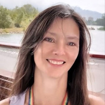 A woman with long brown hair smiles outdoors near water, with trees and mountains in the background.