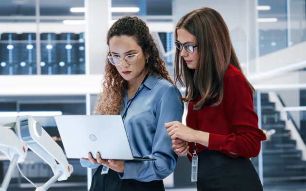 Two women wearing business attire and glasses stand together in a modern office, looking at a laptop screen.