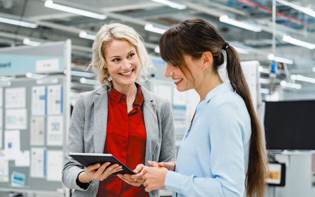 Two women stand in an office or industrial setting, one holding a tablet, both smiling as they have a conversation.