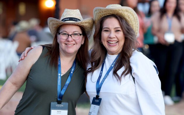 Two women wearing straw hats and conference lanyards smile outdoors, standing close together with one arm around each other.