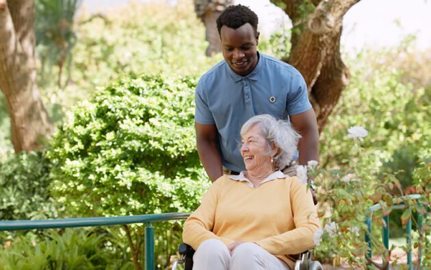 A young man stands behind an older woman in a wheelchair outdoors. Both are smiling, with greenery and trees in the background.