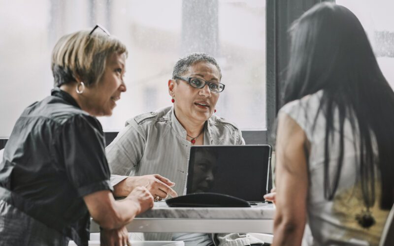 Three women sit around a table having a discussion, with a laptop open in front of them in a well-lit room.