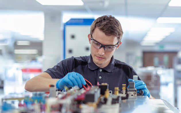 A man wearing safety glasses and gloves works on electronic components at a lab workstation.