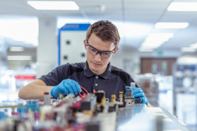 A man wearing safety glasses and gloves works on an electronic circuit board in a well-lit lab or workshop.