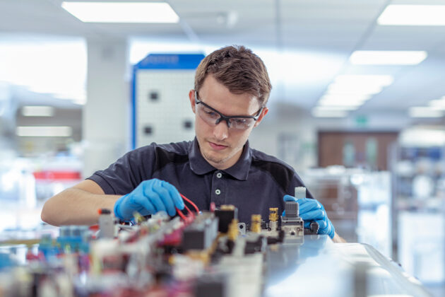 A man wearing safety glasses and gloves works on an electronic circuit board in a well-lit lab or workshop.