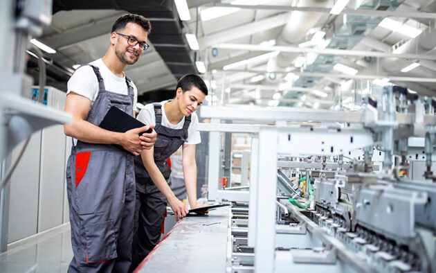 Two workers in gray coveralls operate and inspect machinery in a modern industrial facility. One stands with a tablet, and the other examines equipment on the production line.