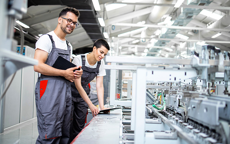 Two factory workers in overalls inspect and monitor automated machinery on a production line inside an industrial facility.