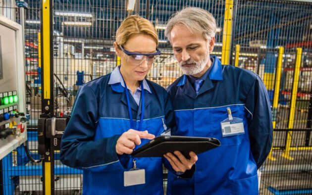 Two factory workers in blue uniforms review data on a tablet while standing in an industrial facility near machinery and safety barriers.