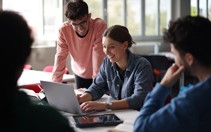 Four young adults sit and stand around a table, collaborating and smiling while looking at a laptop in a well-lit room with large windows.