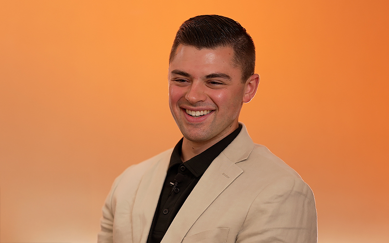 A man wearing a beige blazer over a black shirt smiles in front of a plain orange background.
