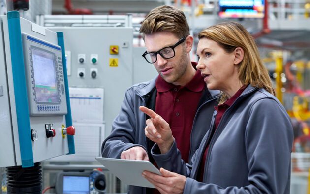 Two people in industrial uniforms review data on a tablet and control panel in a factory setting with machinery in the background.