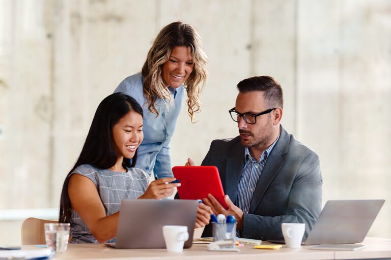 Three colleagues sit at a table with laptops and documents, discussing something on a red tablet. One person stands, smiling, while the other two look at the screen and point.